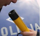Osoba badana na stan trzeźwości  na urządzeniu Alco Blow na tle radiowozu z napisem POLICJA